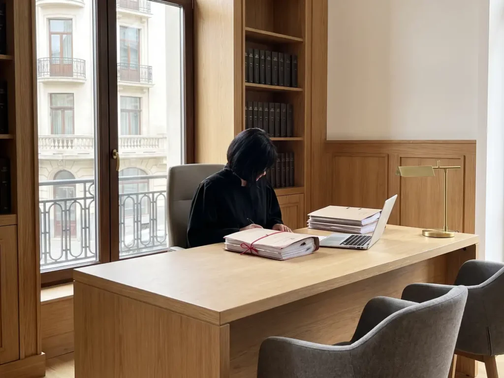 Judecătoare singură în camera de consiliu, cu un dosar legat cu șnur roșu pe birou și scaunele pentru părți goale în fața ei
