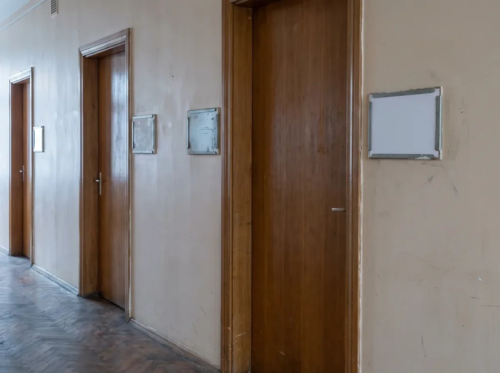 Coridor de institutie cu patru usi inchise; langa trei dintre ele suportul de nume e gol, iar langa a patra sta o fisa alba