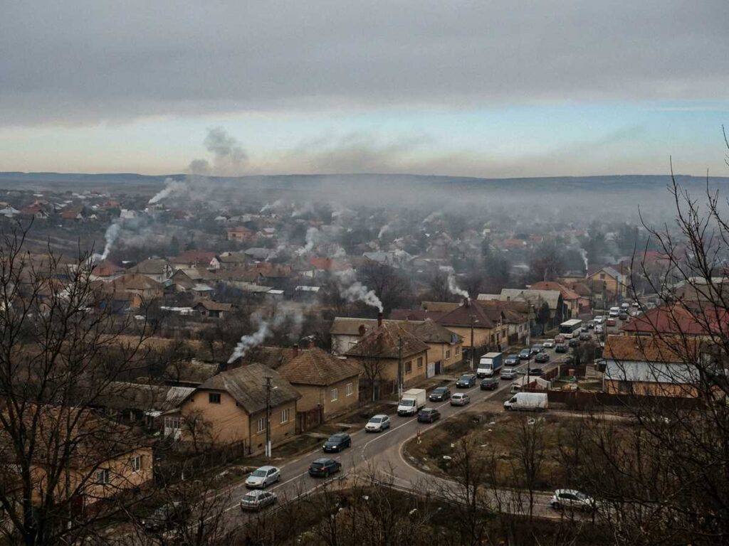 Fum de la sobe peste acoperisurile unui oras romanesc intr-o dimineata rece, cu trafic rutier in fundal