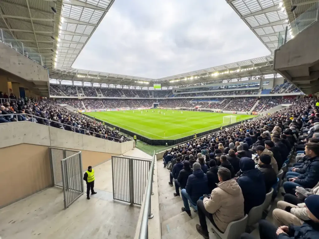 Tribună plină pe un stadion modern din România, cu porțile de evacuare deschise și un agent de ordine lângă ele