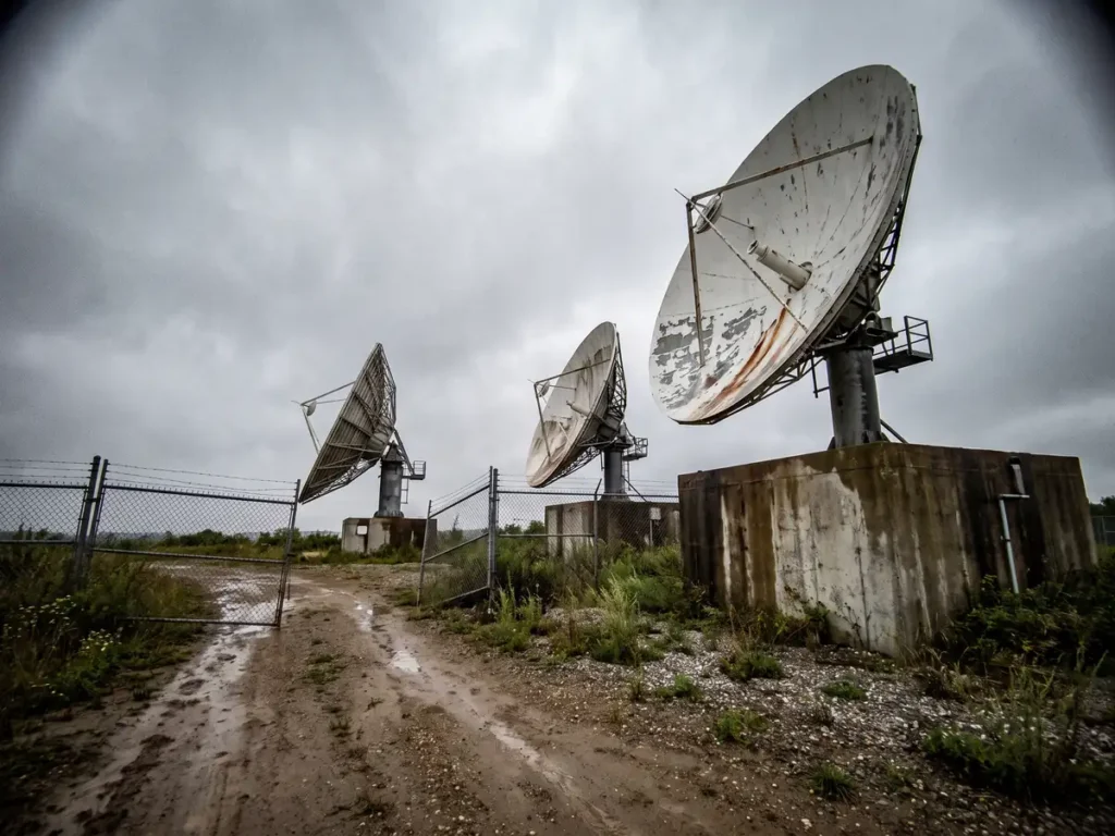 Antene parabolice ruginite ale unei stații terestre de satelit, sub cer înnorat, într-un perimetru împrejmuit cu gard