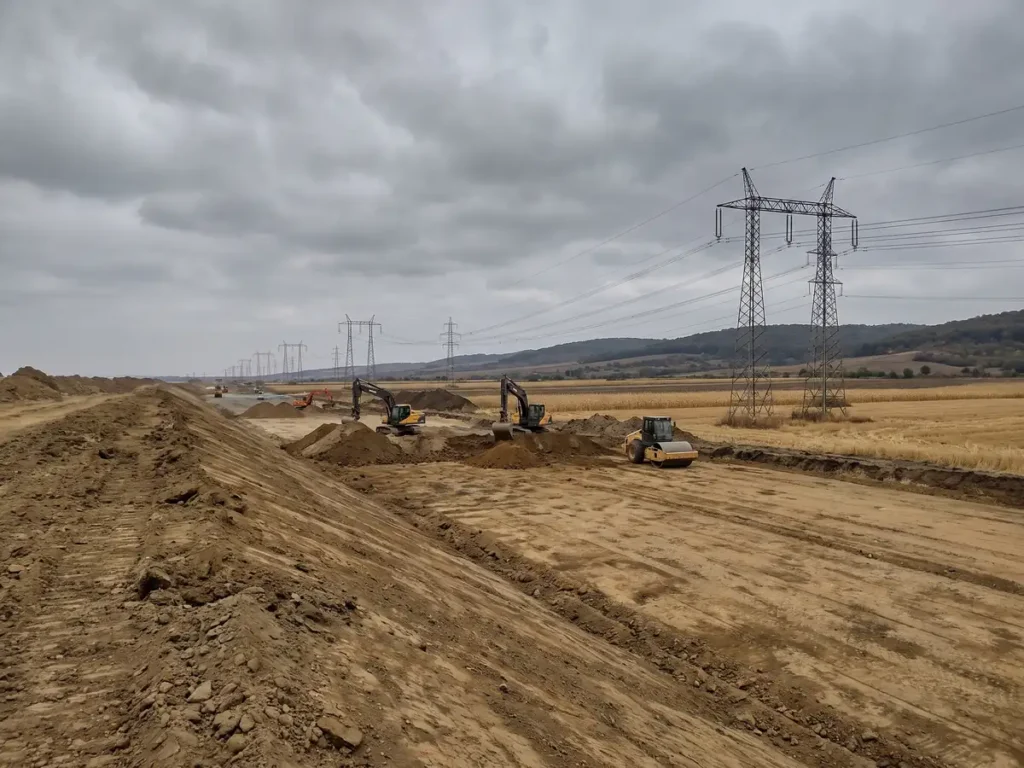 Șantier de autostradă în câmp deschis, cu terasament de pământ, utilaje și stâlpi de electricitate în curs de relocare