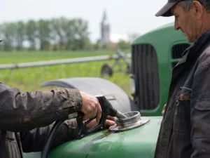 Mainile unui fermier introduc pistolul de motorina in rezervorul unui tractor agricol verde, la marginea unui lan de porumb